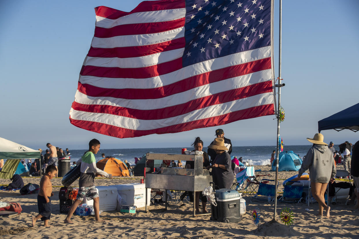 Huntington Beach, CA - July 04: The Espinoza, Vazquez and Romero families of Fallbrook cook tacos on the beach under their giant American flag while waiting for fireworks over the ocean on Sunday, July 4, 2021 in Huntington Beach, CA.  The festival featured live entertainment and DJs, merchant and craft vendor booths, amusement and carnival rides, food trucks, a beer and wine garden, sponsor activations, live radio broadcasts and awards.  A Surf City Run 5K kicked off the day and a neighborhood parade spanning 3 separate routes replaced the grand parade of previous years before the pandemic.  The grand finale featured fireworks over the ocean shot off from the pier at 9 p.m.  (Allen J. Schaben/Los Angeles Times via Getty Images)