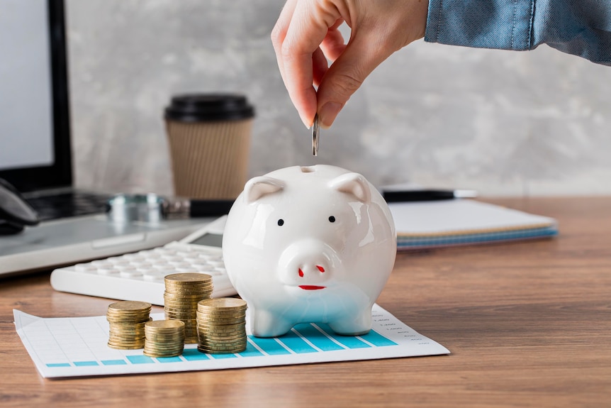 A man about to insert a coin into a white piggy bank with other stacks of coins sitting next to the piggy bank